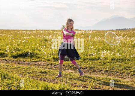 Happy and excited child girl playing in the green meadow lit by the summer sun, making soap bubbles. Childhood innocence concept. Stock Photo