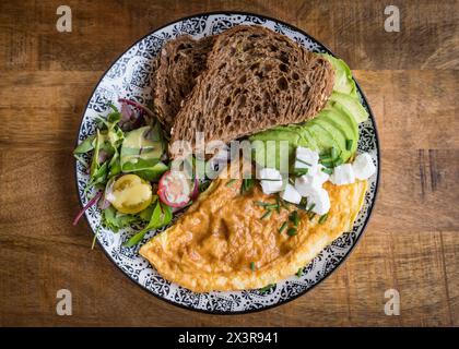 Top view of omelet with feta cheese, avocado, salad and brown bread on a plate on a wooden table in a café restaurant Stock Photo