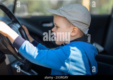 Cute little boy behind the wheel of an fathers car, he holds the steering wheel, happily looks to road. Fulfilling a child's dream of sitting behind t Stock Photo