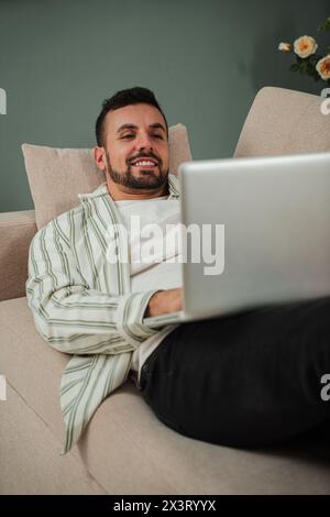 Vertical. Handsome Young Adult Man Using Laptop While Sitting On Couch At Home. Smiley Guy Working Remotely On Computer Laying On A Sofa At Cozy Apartment Living Room. Happy Male Enjoying Online Job. High quality photo Stock Photo