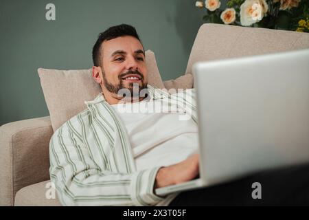 Handsome Young Adult Man Using Laptop While Sitting On Couch At Home. Smiley Millennial Guy Working Remotely On Computer Laying On A Sofa At Cozy Apartment Living Room. Happy Male Enjoying Online Job. High quality photo Stock Photo