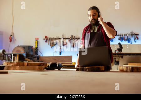 Cabinetmaker receiving telephone call from client inquiring about furniture assembling commission progress. Carpenter in assembly shop talking on smartphone with customer Stock Photo