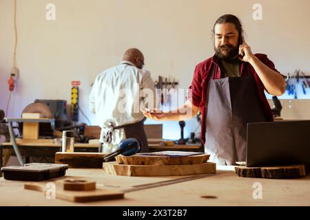 Smiling manufacturer discussing on telephone call with client inquiring about furniture assembling commission progress. Cheerful carpenter in assembly shop talking on mobile phone with customer Stock Photo