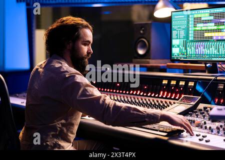 Portrait of music producer working in control room with sliders and switchers, adjusting volume level on audio recordings. Audio engineer works with mixing console in professional studio. Stock Photo