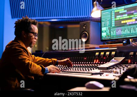 African american sound engineer works on mixing console with sliders and knobs, looking at digital software to record and process tunes. Audio technician creating new music, control room board. Stock Photo