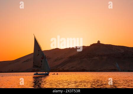 A felucca sailing past the Tombs of the Nobles in Aswan, Egypt at sunset Stock Photo