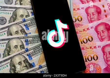 TikTok app logo seen on the screen of smartphone, which is placed on US dollars an Chinese Yuan banknotes. Stafford, United Kingdom, April 28, 2024 Stock Photo