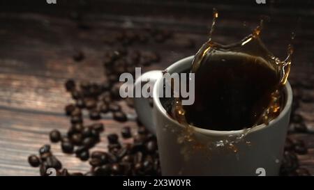Falling drop in hot espresso in coffee cup decorated with piles of coffee bean placed on wooden table surrounded with black background. Close up of Stock Photo