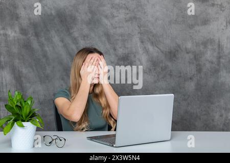 Young lady with blonde hair in gray dress posing near laptop and green flower Stock Photo