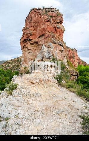 A rust-red and cream sandstone rock tower rises above a pale rocky slope with green shrubs at Ayres Natural Bridge Park, Wyoming. Stock Photo