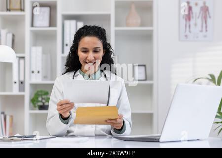 A cheerful female doctor in her office, smiling as she examines patient documents. Her workstation is tidy with a laptop open. Represents dedication and care in healthcare. Stock Photo