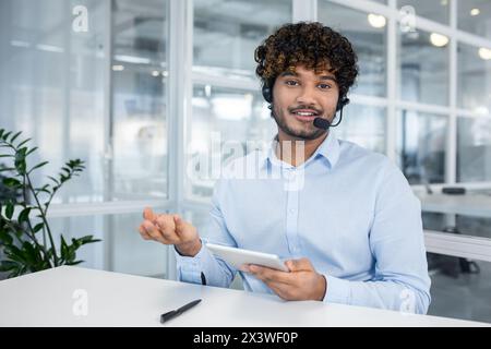 Young man in a light blue shirt and headset smiles engagingly as he talks over a video call, using a tablet. Modern office setting with glass walls. Stock Photo