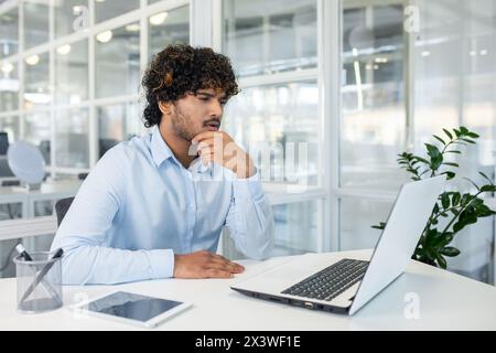 A young professional man is deeply focused as he analyzes data on a laptop within a bright, modern office setting. Conceptualizing business strategies. Stock Photo