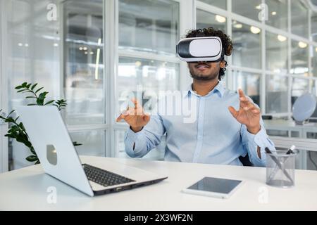 Curly-haired young professional immersed in virtual reality using a VR headset while engaging with invisible interfaces at his workplace equipped with various modern devices. Stock Photo