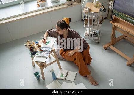 Top view of focused woman artist sitting on floor in art studio painting watercolours in sketchbook  Stock Photo