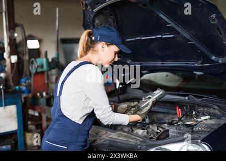 Female auto mechanic using diagnostic tool, scanner, running diagnostic on car. Beautiful woman working in a garage, wearing blue coveralls. Stock Photo