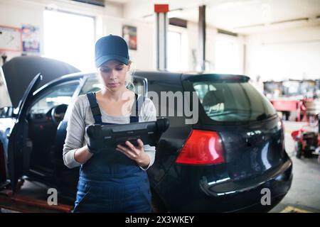 Female auto mechanic using diagnostic tool, scanner, running diagnostic on car. Beautiful woman working in a garage, wearing blue coveralls. Stock Photo