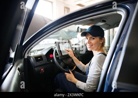 Female auto mechanic using diagnostic tool, scanner, running diagnostic on car. Beautiful woman sitting inside of car, performing diagnostic test. Stock Photo
