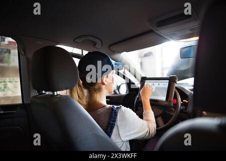 Female auto mechanic using diagnostic tool, scanner, running diagnostic on car. Beautiful woman sitting inside of car, performing diagnostic test. Stock Photo