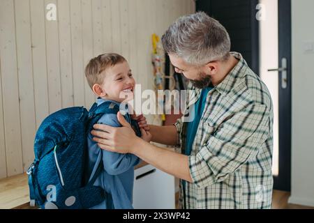 Father helping son get ready for kindergarten, preschool. Putting backpack on back. Stock Photo