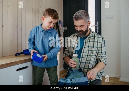 Father helping son get ready for kindergarten, preschool. Putting lunchbox with food, snacks in backpack. Stock Photo