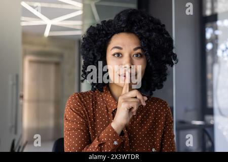 African American woman with curly hair making a shushing gesture, signaling silence in a contemporary office environment. Her playful expression conveys secrecy or surprise. Stock Photo