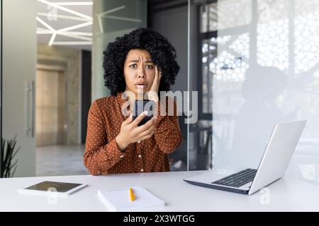 African American businesswoman in a modern office, looking shocked while reading a message on her smartphone. Laptop and documents on her desk. Stock Photo