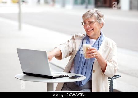 Happy smiling Elderly Senior Business woman with gray short hair, Mature female freelancer works on laptop, looks at computer at table in outdoor cafe Stock Photo