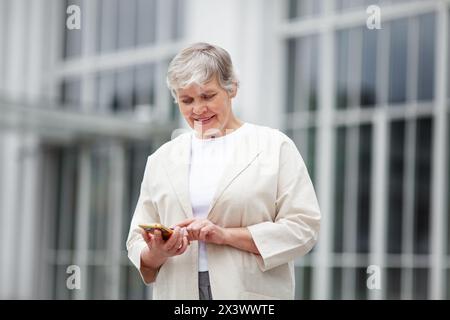 Mature beautiful woman with smartphone, positive model standing outdoor on street, dressed in light clothing. Elderly female senior with gray hair on Stock Photo