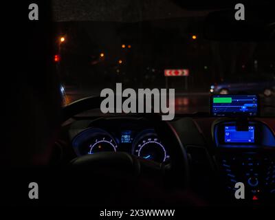 Driver sits behind the wheel and drives in a car through dark city at night. Raindrops on a windshield, highlighted dashboard, road traffic. Stock Photo