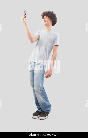 Young happy smiling handsome man using his smartphone, taking a selfie isolated against a gray background, full body image Stock Photo