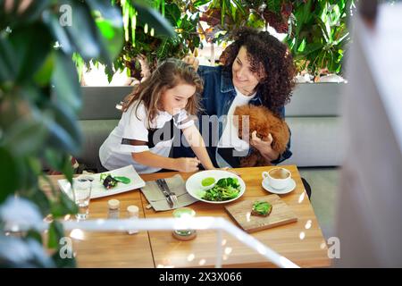 A woman and a girl are seated at a table, enjoying vegetables meal together. Various dishes and utensils are spread out on the table as they engage in Stock Photo