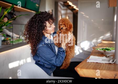 Portrait of a beautiful curly brunette woman cuddling her poodle pet in a restaurant. Elegant female with her small dog enjoying indoor dining. Stock Photo