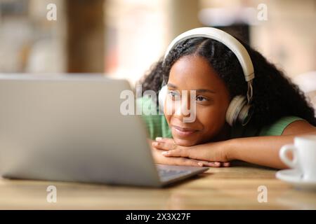 Satisfied black woman with headphone watching media content on laptop in a restaurant terrace Stock Photo