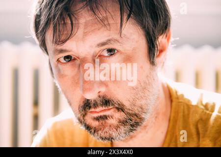 Portrait of mid adult unkempt male with beard in late 40s looking at camera in his living room back lit by the morning sun Stock Photo