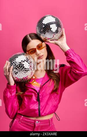 A stylish woman in her 20s, clad in a pink jacket, holds two disco balls in a studio with a vibrant pink background. Stock Photo