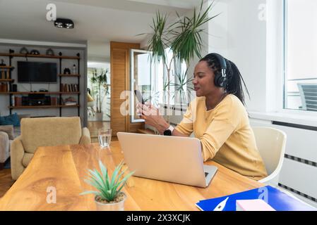 Happy woman wearing headphones sitting with laptop at table in home office. Successful African American freelance business female working online liste Stock Photo