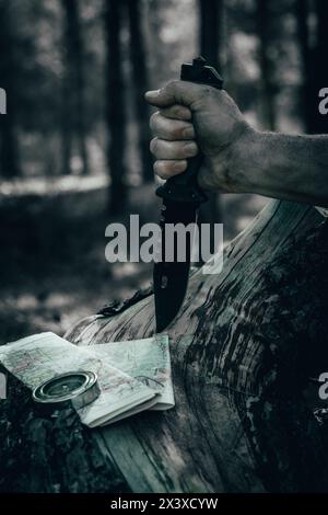 Man hand in forest holds hunting knife against background terrain map and compass. Concept survival wilderness Stock Photo