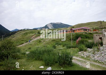 A small wooden home with a red roof on a hill, overlooking a valley and mountains. Blue sky, white clouds, green grass. Stock Photo