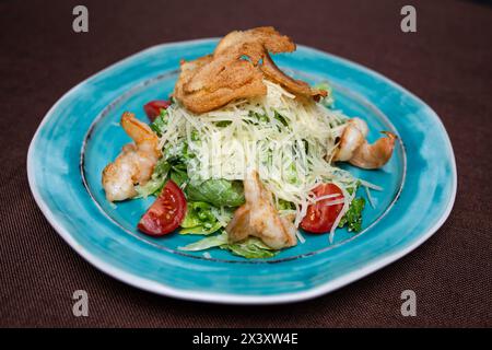 Healthy and delicious caesar salad with fresh greens, juicy tomatoes, and tasty shrimps on a blue plate. Isolated on a brown background. Stock Photo
