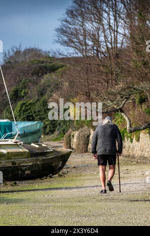 A mature man with a walking stick walking past various sailing craft boats sail boats in various states of disrepair moored at low tide on the tidal G Stock Photo