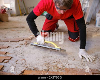 Craftsman in workwear who, using the rabot, removes excess glue left on the floor after removing the wooden parquet glued to the slab of an apartment. Stock Photo