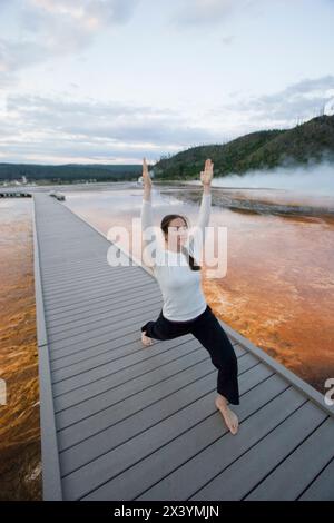 As the sun rises, a woman practices yoga on a boardwalk over algae beds as steam rises from the Grand Prismatic Spring in Yellow Stock Photo