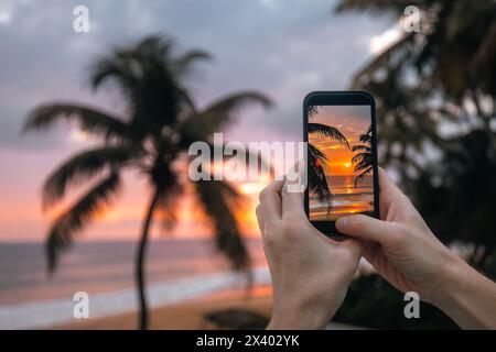 Close-up of hands holding smart phone. Man photographing sunset over ocean with mobile phone. Coconut palm trees on sand beach in south coast of Sri L Stock Photo