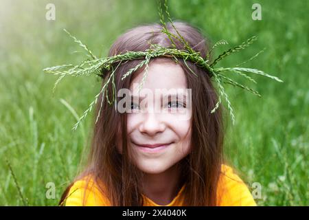 A little girl in a wreath of spikelets of grass against a background of blurred greenery. Stock Photo