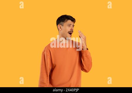 Young Man in Orange Sweatshirt Yawning Against Bright Yellow Background Stock Photo