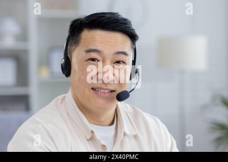 Portrait of a happy middle-aged Asian man wearing a headset, working remotely from his home office. He shows a friendly and professional attitude. Stock Photo