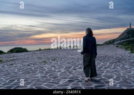 Woman in a long coat stands looking out over the Mediterranean Sea at sunset, with Capo Sandalo Lighthouse in the background, on San Pietro Island ... Stock Photo