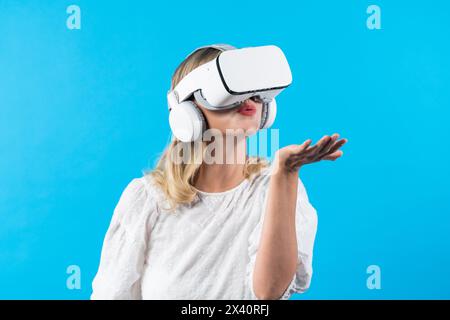Caucasian girl with VR goggles and standing while holding something and standing at blue background. Woman blowing and looking at hologram in hands Stock Photo