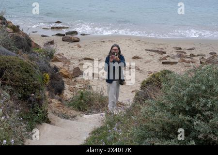 Woman standing on steps on a beach takes a photograph with a smart phone camera towards the photographer's camera; Mykonos, Greece Stock Photo